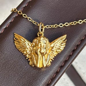 May include: A gold-colored pendant necklace featuring a detailed angel with outstretched wings. The angel pendant is suspended from a delicate gold chain. The necklace is displayed on a brown leather surface.