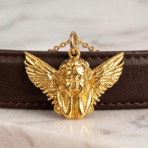 May include: A gold-colored cherub pendant necklace. The cherub has detailed wings and is suspended from a delicate chain. The necklace is displayed against a brown leather surface and a white marble background.