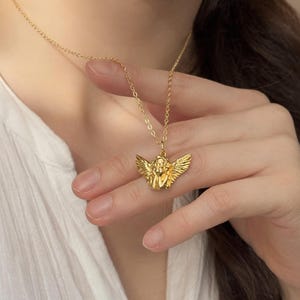 May include: Gold-colored necklace featuring a pendant of an angel with outstretched wings. The angel pendant is intricately detailed and suspended from a delicate chain. The necklace is worn against a white shirt.