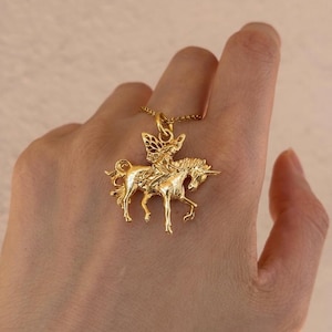 May include: A gold-toned metal pendant necklace featuring a detailed relief of a winged fairy figure riding a unicorn. The pendant is suspended from a delicate gold-toned chain, showcasing intricate textures on the mythical creature and rider.