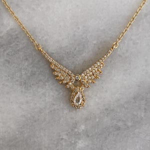Puede incluir: Un collar dorado con un colgante en forma de alas de &aacute;ngel. Las alas y un colgante en forma de l&aacute;grima est&aacute;n engastados con peque&ntilde;os cristales transparentes. La cadena es delicada y tambi&eacute;n dorada.