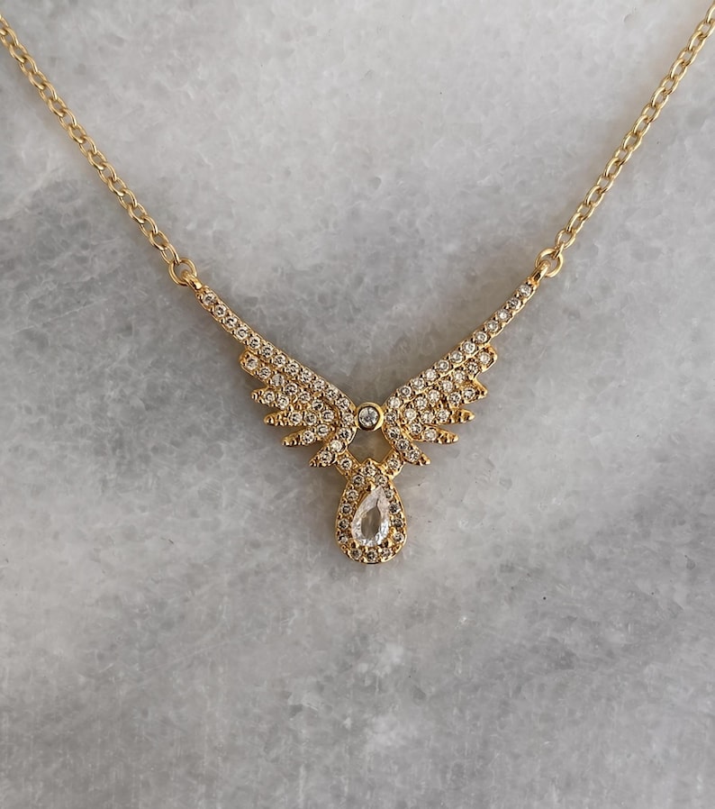 Puede incluir: Un collar dorado con un colgante en forma de alas de &aacute;ngel. Las alas y un colgante en forma de l&aacute;grima est&aacute;n engastados con peque&ntilde;os cristales transparentes. La cadena es delicada y tambi&eacute;n dorada.