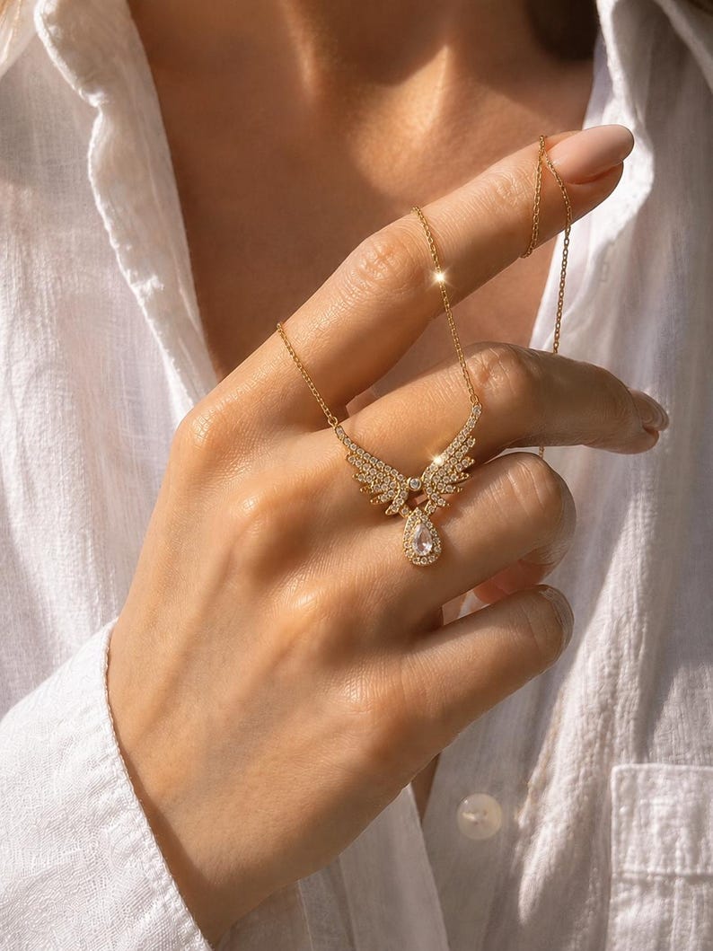 Puede incluir: Un collar dorado con un colgante en forma de alas de &aacute;ngel, adornado con peque&ntilde;os cristales brillantes. El colgante tiene una gema en forma de l&aacute;grima en el centro. El collar se muestra sobre una camisa blanca.