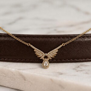 Puede incluir: Un collar dorado con un colgante en forma de ala, incrustado con peque&ntilde;as piedras preciosas transparentes. El colgante presenta una gema en forma de l&aacute;grima en la parte inferior. El collar se exhibe sobre una superficie marr&oacute;n sobre un fondo de m&aacute;rmol.