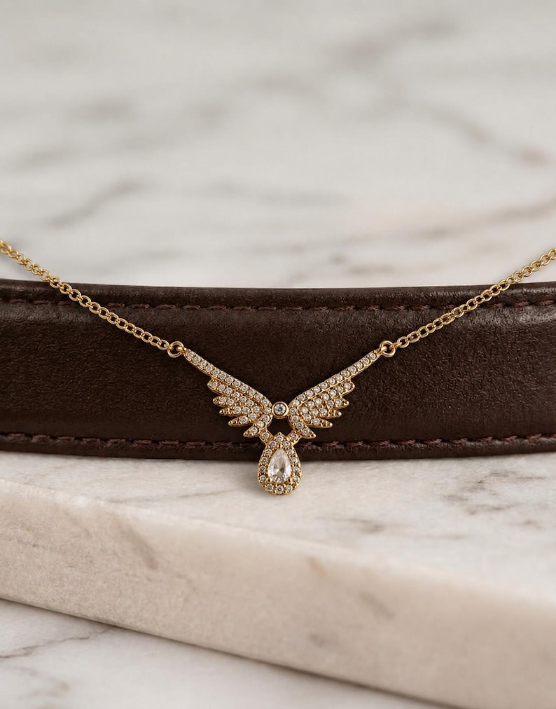 Puede incluir: Un collar dorado con un colgante en forma de ala, incrustado con peque&ntilde;as piedras preciosas transparentes. El colgante presenta una gema en forma de l&aacute;grima en la parte inferior. El collar se exhibe sobre una superficie marr&oacute;n sobre un fondo de m&aacute;rmol.