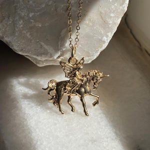 May include: A gold-toned necklace featuring a pendant of a winged fairy riding a unicorn. The pendant is suspended from a delicate gold-toned chain. The design shows the fairy with wings seated on the back of a unicorn with a single horn.