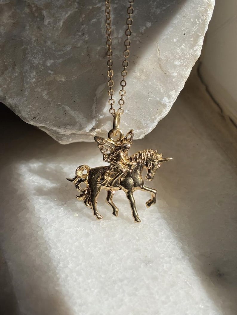 May include: A gold-toned necklace featuring a pendant of a winged fairy riding a unicorn. The pendant is suspended from a delicate gold-toned chain. The design shows the fairy with wings seated on the back of a unicorn with a single horn.
