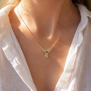 Puede incluir: Un collar dorado con un colgante en forma de alas adornado con peque&ntilde;as piedras brillantes y una gema central en forma de l&aacute;grima. El collar se muestra sobre una camisa blanca.