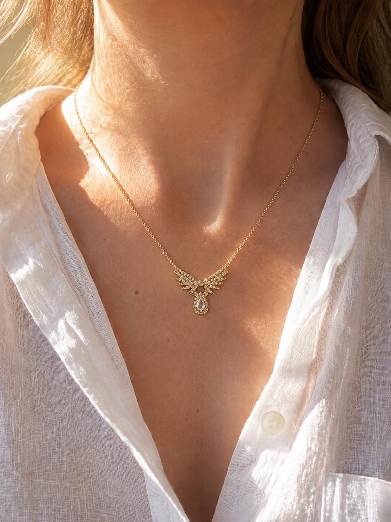 Puede incluir: Un collar dorado con un colgante en forma de alas adornado con peque&ntilde;as piedras brillantes y una gema central en forma de l&aacute;grima. El collar se muestra sobre una camisa blanca.