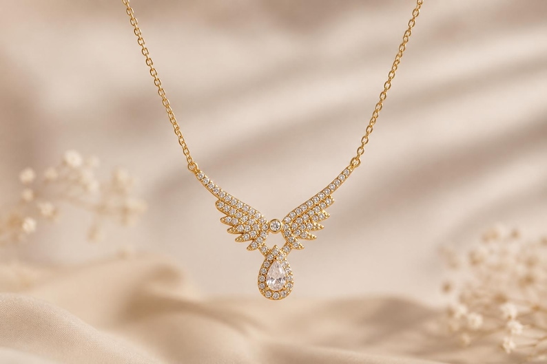 Puede incluir: Un collar dorado con un colgante en forma de alas de &aacute;ngel y una l&aacute;grima. Las alas y la l&aacute;grima est&aacute;n incrustadas con peque&ntilde;os cristales brillantes. El collar cuelga de una delicada cadena.