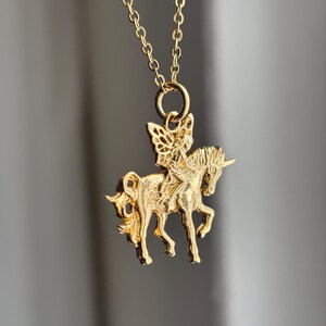 May include: A gold-toned necklace featuring a pendant shaped like a fairy riding a unicorn. The pendant is suspended from a delicate gold-toned chain link. The design shows the fairy with wings seated on the back of the unicorn, which has a horn.