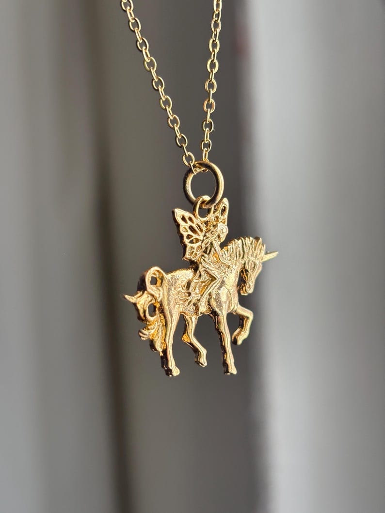 May include: A gold-toned necklace featuring a pendant shaped like a fairy riding a unicorn. The pendant is suspended from a delicate gold-toned chain link. The design shows the fairy with wings seated on the back of the unicorn, which has a horn.