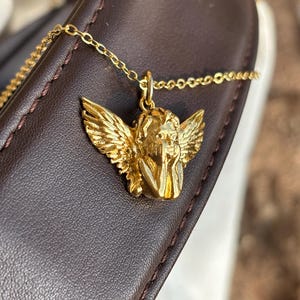 May include: A gold-colored necklace featuring a pendant of a cherub with detailed wings. The cherub is resting its head on its hand. The necklace chain is delicate and gold-toned. The pendant is set against a dark brown leather surface.