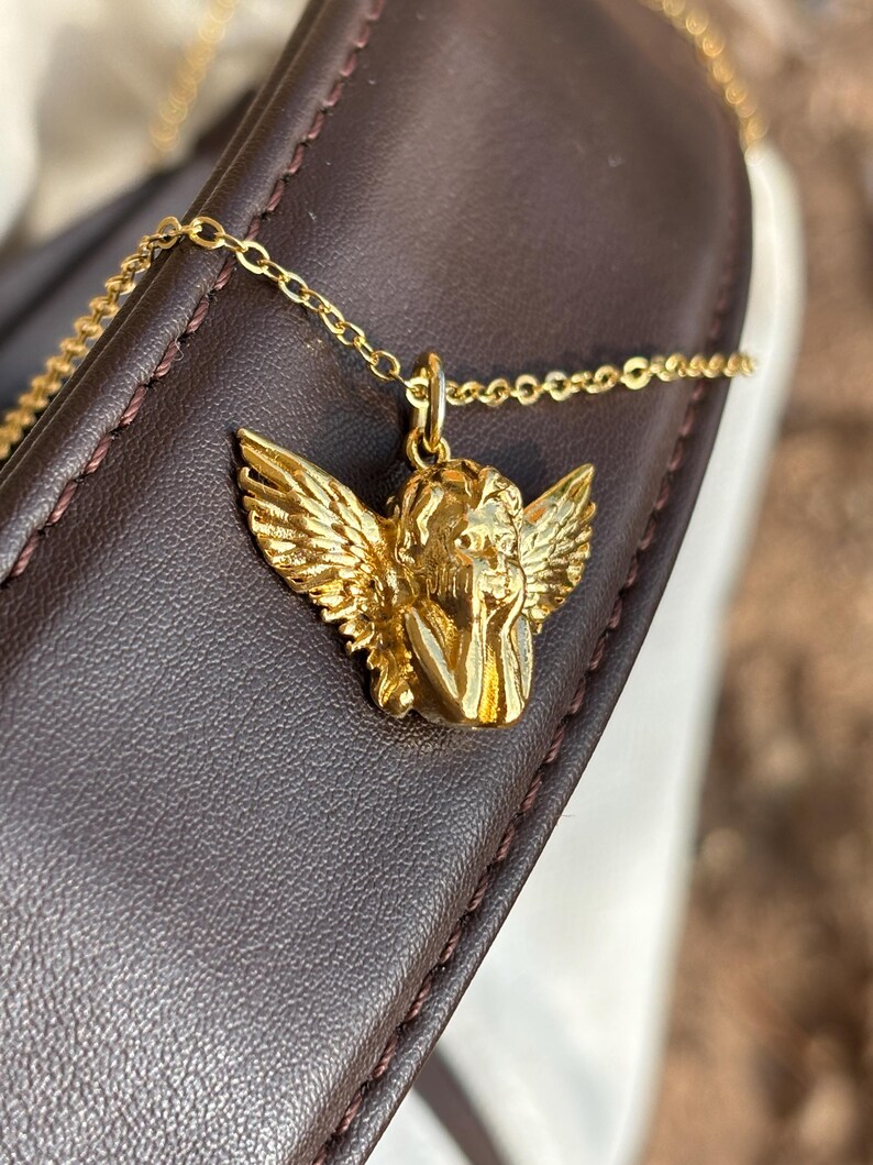 May include: A gold-colored necklace featuring a pendant of a cherub with detailed wings. The cherub is resting its head on its hand. The necklace chain is delicate and gold-toned. The pendant is set against a dark brown leather surface.