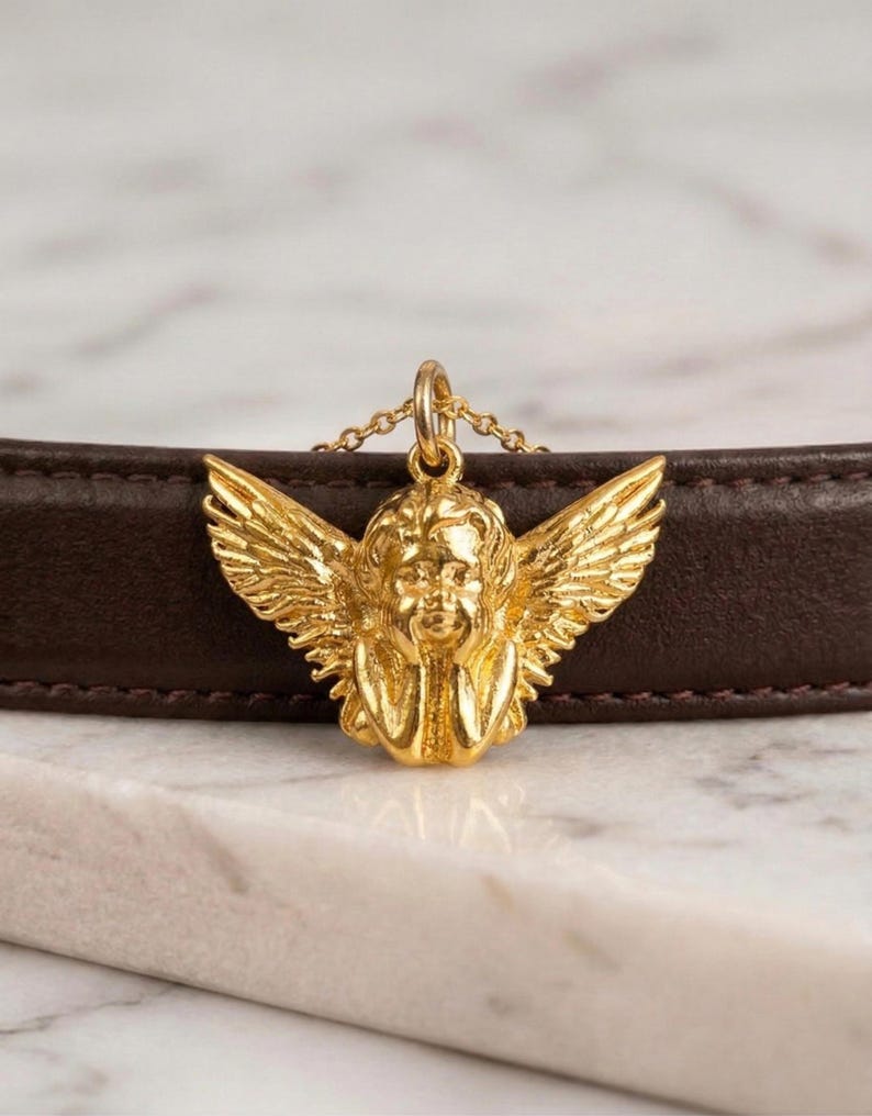 May include: A gold-colored cherub pendant necklace. The cherub has detailed wings and is suspended from a delicate chain. The necklace is displayed against a brown leather surface and a white marble background.