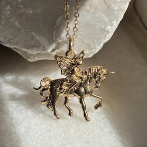 May include: A gold-toned metal pendant necklace featuring a detailed figure of a fairy riding a unicorn. The pendant is suspended from a delicate gold-toned chain, showcasing the intricate sculpted design of the mythical creature and rider.