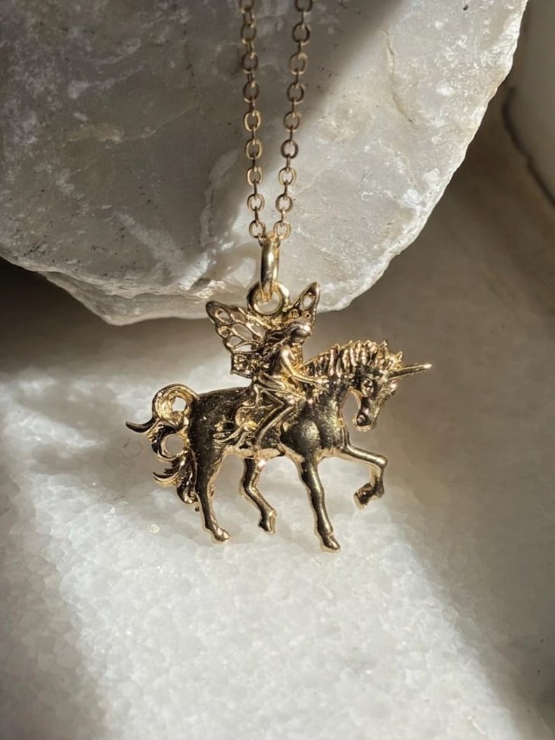 May include: A gold-toned metal pendant necklace featuring a detailed figure of a fairy riding a unicorn. The pendant is suspended from a delicate gold-toned chain, showcasing the intricate sculpted design of the mythical creature and rider.
