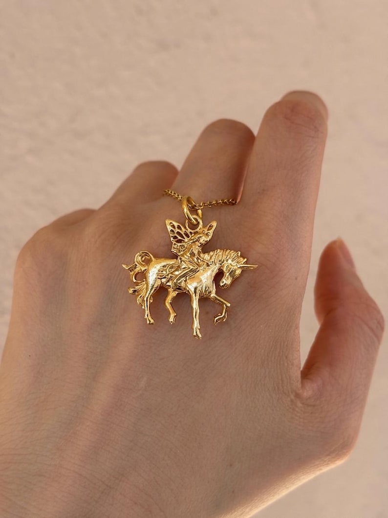 May include: A gold-toned metal pendant necklace featuring a detailed relief of a winged fairy figure riding a unicorn. The pendant is suspended from a delicate gold-toned chain, showcasing intricate textures on the mythical creature and rider.