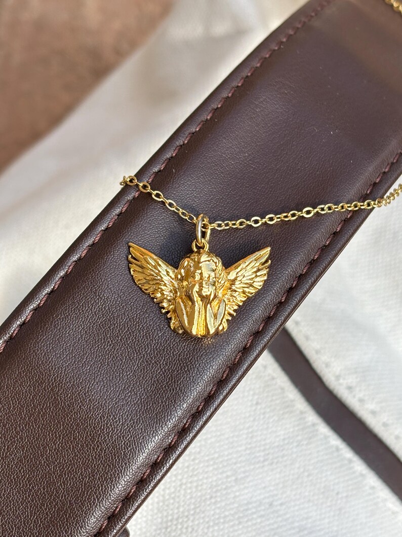 May include: A gold-colored pendant necklace featuring a detailed angel with outstretched wings. The angel pendant is suspended from a delicate gold chain. The necklace is displayed on a brown leather surface.