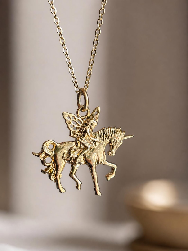 May include: A gold-toned metal pendant necklace featuring a detailed figure with wings riding a unicorn. The pendant is suspended from a delicate gold-toned chain. The design shows the winged figure seated on the back of the unicorn, which is depicted in a standing position with its horn clearly visible.