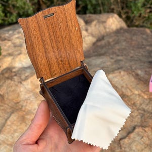 May include: A small, open wooden box with a dark brown finish and a hinged lid. The interior of the box is lined with black fabric. A white, rectangular cleaning cloth with pinked edges is draped over the side of the open box.