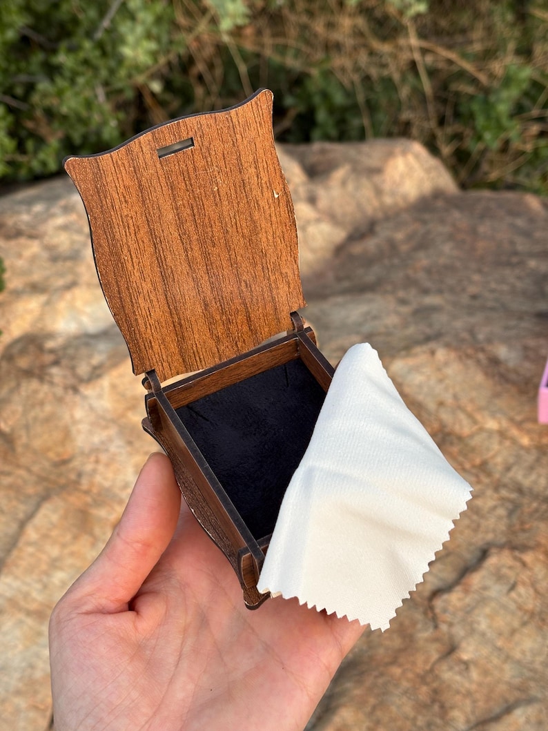 May include: A small, open wooden box with a dark brown finish and a hinged lid. The interior of the box is lined with black fabric. A white, rectangular cleaning cloth with pinked edges is draped over the side of the open box.