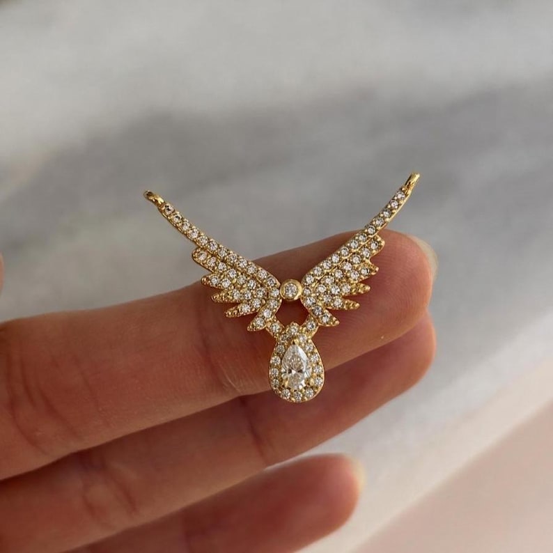 Puede incluir: Un colgante dorado con forma de alas de &aacute;ngel, incrustado con peque&ntilde;as piedras preciosas transparentes. El colgante presenta una piedra preciosa en forma de pera en la parte inferior. Las alas est&aacute;n extendidas, creando un dise&ntilde;o sim&eacute;trico.