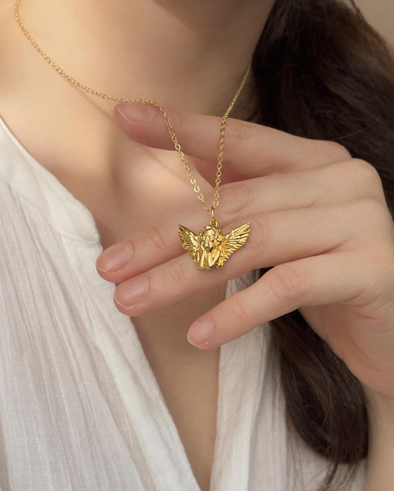 May include: Gold-colored necklace featuring a pendant of an angel with outstretched wings. The angel pendant is intricately detailed and suspended from a delicate chain. The necklace is worn against a white shirt.