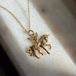 May include: A gold-toned metal necklace featuring a pendant of a winged figure riding a unicorn. The pendant is suspended from a delicate gold-toned chain. The design shows the unicorn in a prancing pose with the winged figure seated on its back.