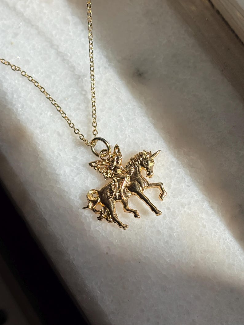 May include: A gold-toned metal necklace featuring a pendant of a winged figure riding a unicorn. The pendant is suspended from a delicate gold-toned chain. The design shows the unicorn in a prancing pose with the winged figure seated on its back.
