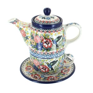 May include: A ceramic teapot and teacup set with a floral and hummingbird design. The set includes a teapot with a lid, a teacup, and a saucer. The design features red, blue, and yellow flowers with hummingbirds on a white background.