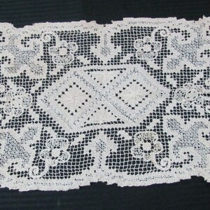 May include: Cream-colored, rectangular lace doily with a floral and geometric pattern. The doily features a central diamond design with floral accents and a scalloped edge. The lace is intricately woven.