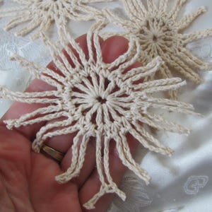 May include: Close-up of several cream-colored, handmade crochet doilies. The doilies have a sunburst design with a central circle and radiating points. The texture of the crochet work is visible, and the doilies are on a white background.