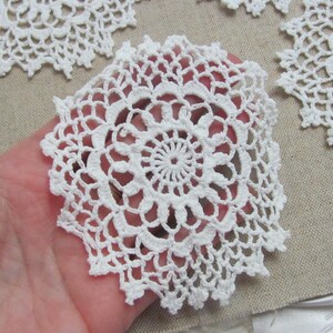 May include: Close-up of several white, crocheted doilies. The doilies have a circular design with intricate openwork patterns. One doily is held in a hand, while others are arranged on a neutral-colored fabric background. The doilies are made of a soft, white yarn.