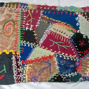 May include: A rectangular textile piece featuring a patchwork of various fabric scraps in shades of brown, burgundy, blue, and black. The segments are joined by decorative, colourful embroidery stitches in yellow, red, green, and white thread.