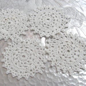 Puede incluir: Cuatro posavasos de ganchillo de color crema. Cada posavasos tiene un centro circular con un patrón calado decorativo y un borde festoneado. Los posavasos están dispuestos sobre una superficie clara y texturizada.