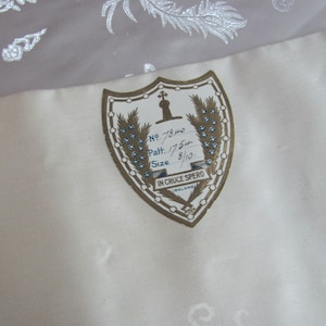 May include: A white fabric label with a gold shield design. The shield has a cross and a sword at the top, and the text "IN CRUCE SPERO" at the bottom. The label also has the text "No. 7840, Patt. 1754, Size 8/10."