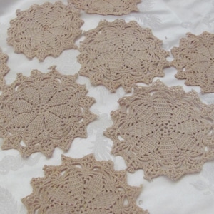Puede incluir: Una colección de tapetes de ganchillo beige dispuestos sobre una superficie blanca. Los tapetes tienen forma circular con intrincados patrones calados y bordes festoneados. Probablemente hechos a mano, añaden un toque vintage a la decoración del hogar.