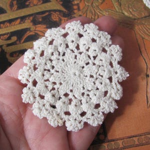 May include: A small, circular, off-white crochet doily featuring an intricate, repeating floral-inspired pattern with a scalloped edge. The delicate textile piece is crafted from fine thread, showcasing a detailed openwork design throughout the center.