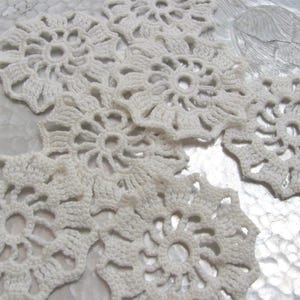Puede incluir: Primer plano de varios tapetes de ganchillo de color crema. Cada tapete presenta un diseño floral con un agujero central y bordes intrincados en forma de pétalos. Los tapetes están dispuestos sobre una superficie texturizada y de color claro, creando una estética vintage.