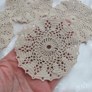 May include: Several beige crocheted doilies with a circular design are displayed. The doilies have a central hole and intricate openwork patterns. The image shows the doilies in a close-up view, highlighting the texture and detail of the handmade craft.
