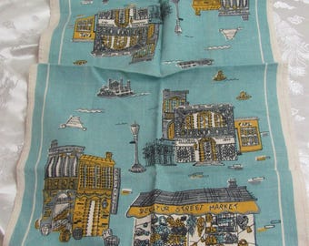 Puede incluir: Un paño de cocina de lino turquesa vintage con ilustraciones de un mercado y edificios de la ciudad. El diseño incluye detalles amarillos, grises y blancos, con el texto "Decatur Street Market" y "Suzanne Meister."