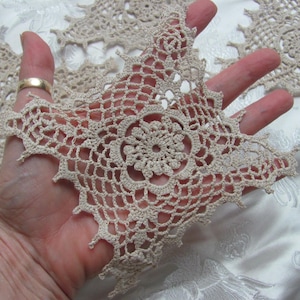 May include: A beige, handmade crochet doily with a floral center and a starburst design. The doily is held in a hand, showcasing its intricate openwork pattern. The delicate lace-like texture is visible, highlighting the craftsmanship of the piece.
