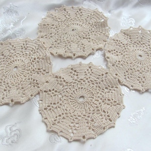 Puede incluir: Cuatro posavasos de ganchillo color crema dispuestos sobre una superficie blanca estampada. Cada posavasos tiene forma circular con un agujero central y patrones calados intrincados. Los posavasos son hechos a mano.