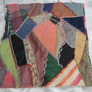 May include: A patchwork textile featuring a variety of fabric scraps in shades of pink, green, blue, and brown. The pieces are sewn together with decorative stitching, creating a textured and colorful design. The textile is square-shaped and appears to be handmade.