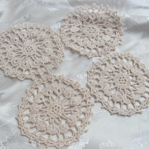 Puede incluir: Cuatro posavasos de ganchillo color crema dispuestos sobre una superficie blanca estampada. Cada posavasos presenta un diseño floral circular con detalles intrincados. Los posavasos son hechos a mano.