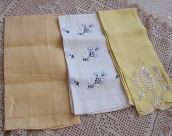 Puede incluir: Tres toallas de mano bordadas. Una es blanca con un diseño floral, otra es amarilla con un diseño floral y la última es amarilla con un borde festoneado.