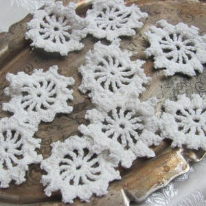 Puede incluir: Una colección de copos de nieve blancos de ganchillo dispuestos sobre una bandeja vintage de tono plateado. Cada copo de nieve presenta un diseño circular detallado con puntas radiantes. La bandeja tiene una apariencia envejecida y deslustrada.