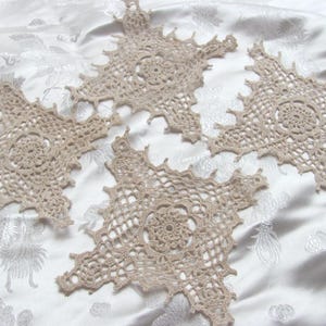Puede incluir: Cuatro tapetes de ganchillo beige en forma de estrella. Cada tapete presenta un diseño floral central y un delicado patrón calado. Los tapetes están dispuestos sobre una tela blanca estampada, mostrando sus intrincados detalles.