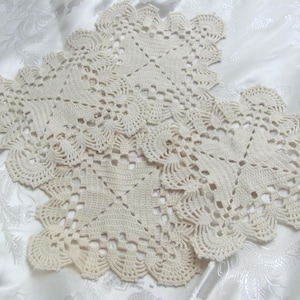 Puede incluir: Cuatro tapetes de ganchillo color crema dispuestos sobre una tela blanca estampada. Cada tapete presenta un diseño central en forma de estrella con calados y bordes festoneados. Estas piezas hechas a mano son probablemente decorativas.